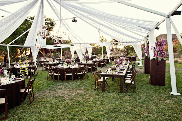 Outdoor reception seating under white tent with chandeliers and purple floral arrangements - photo by Orange County based wedding photographers Mark Brooke